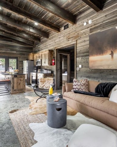 Open-plan cabin interior with weathered wood beams, tan sofa, fishing artwork on wall, and kitchen area visible in background