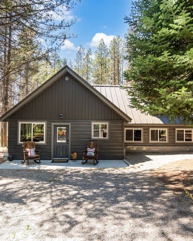 Modern dark wood cabin with metal roof surrounded by tall pine trees and gravel driveway