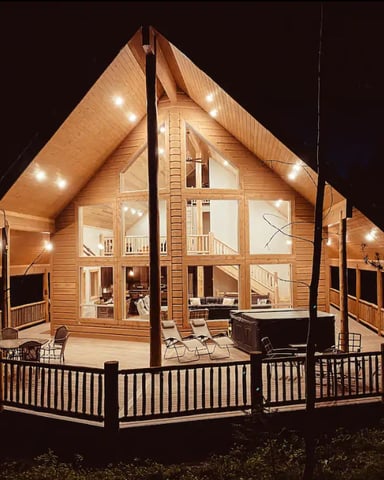 Modern A-frame cabin with wood siding, large windows, and wraparound deck lit up at night with hot tub visible