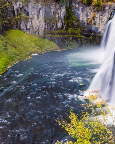 Waterfall cascading into a rocky river gorge surrounded by moss-covered cliffs and autumn foliage