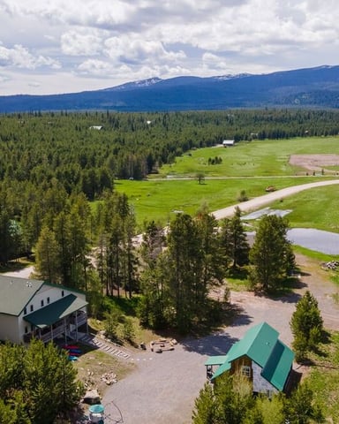 Aerial view of rural property with two houses surrounded by coniferous forest, green meadows, ponds, and snow-capped mountain in distance
