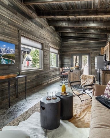 Modern rustic cabin interior with weathered wood walls, exposed beams, stone fireplace, and seating area with mountain lake view on television.