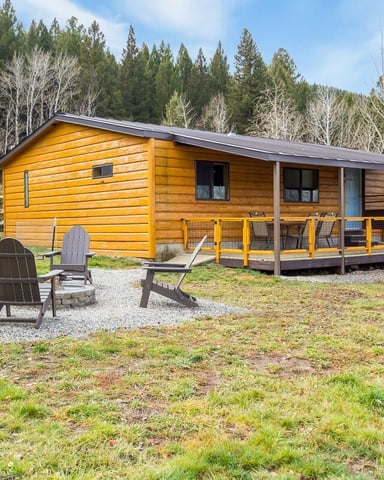 Yellow log cabin with covered porch and yellow railing surrounded by forest, Adirondack chairs in gravel fire pit area.