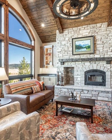 Open-plan living room with stone fireplace, wood ceiling, leather seating, and expansive windows overlooking valley landscape.
