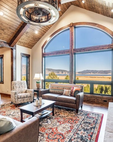Spacious living room with wood ceiling, large arched windows overlooking mountains and lake, fireplace, and mixed seating.