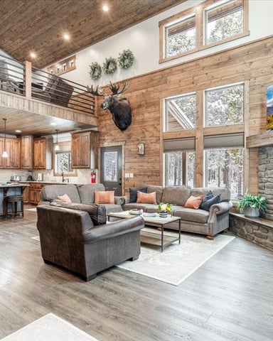 Open-plan cabin interior with wood beams, stone fireplace, gray seating area, and loft mezzanine above kitchen.
