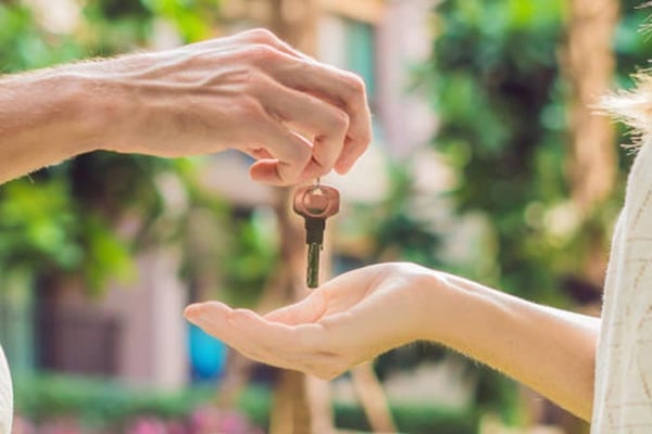 One hand passing a house key to another hand outdoors with blurred residential building and greenery in background
