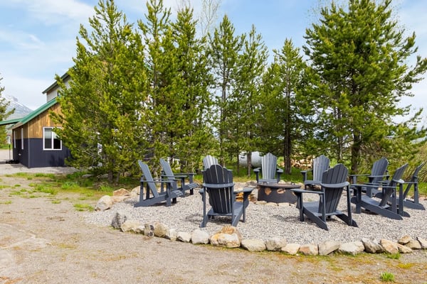 Cabin with green roof surrounded by tall pine trees, fire pit area with dark Adirondack chairs on gravel ground