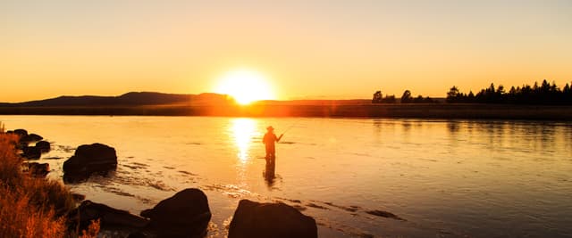 Person fly fishing in calm lake at sunset with forested shoreline and mountains in background