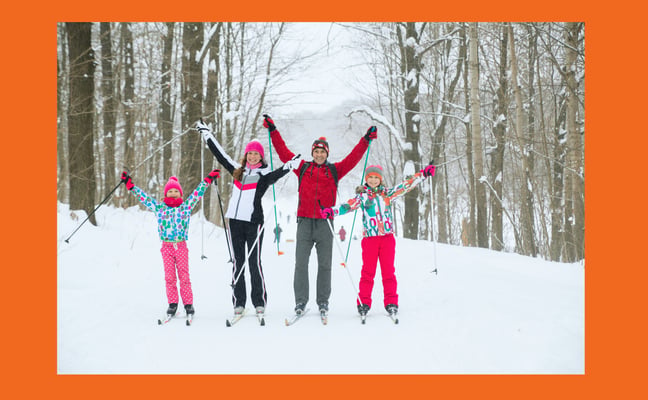 Four children cross-country skiing in snowy forest with arms raised in celebration