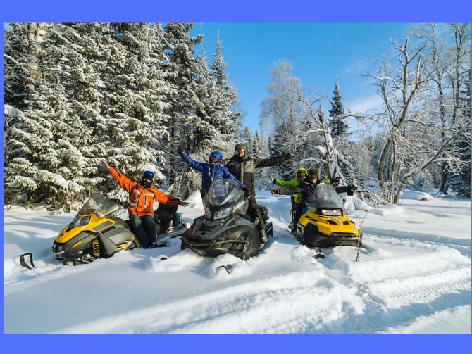 Group of people celebrating on yellow and black snowmobiles in snowy forest with evergreen and birch trees under clear blue sky